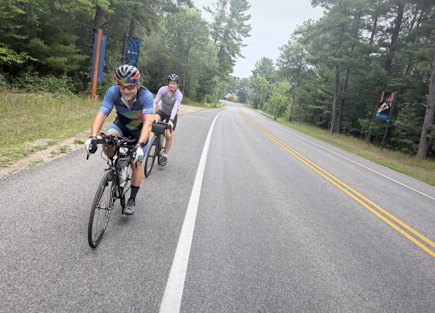 Two Pedal for Pride cyclists riding together on a wooded Michigan road during the 2026 ride.