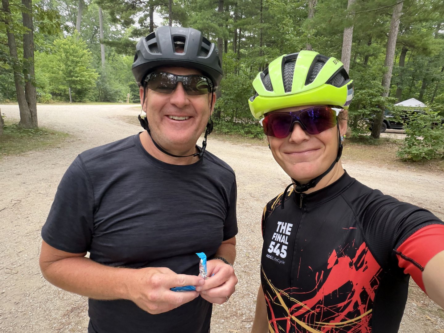 Two Pedal for Pride riders smiling for a photograph during a wooded Day 1 stop.