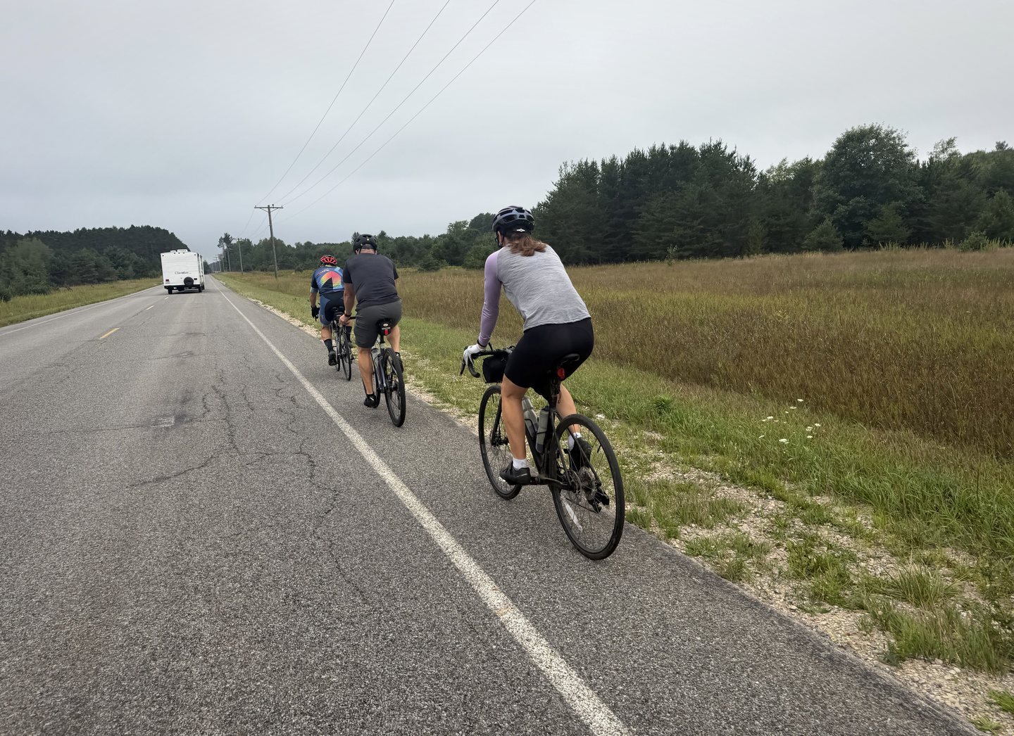 Three cyclists riding along the shoulder of a quiet country road.