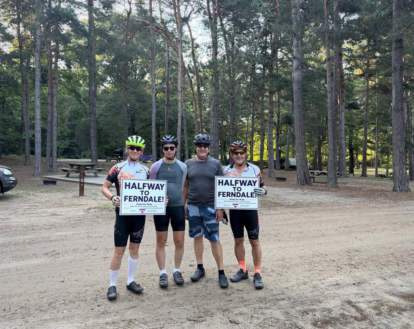 Four Pedal for Pride riders holding Halfway to Ferndale signs at a wooded rest stop in 2026.