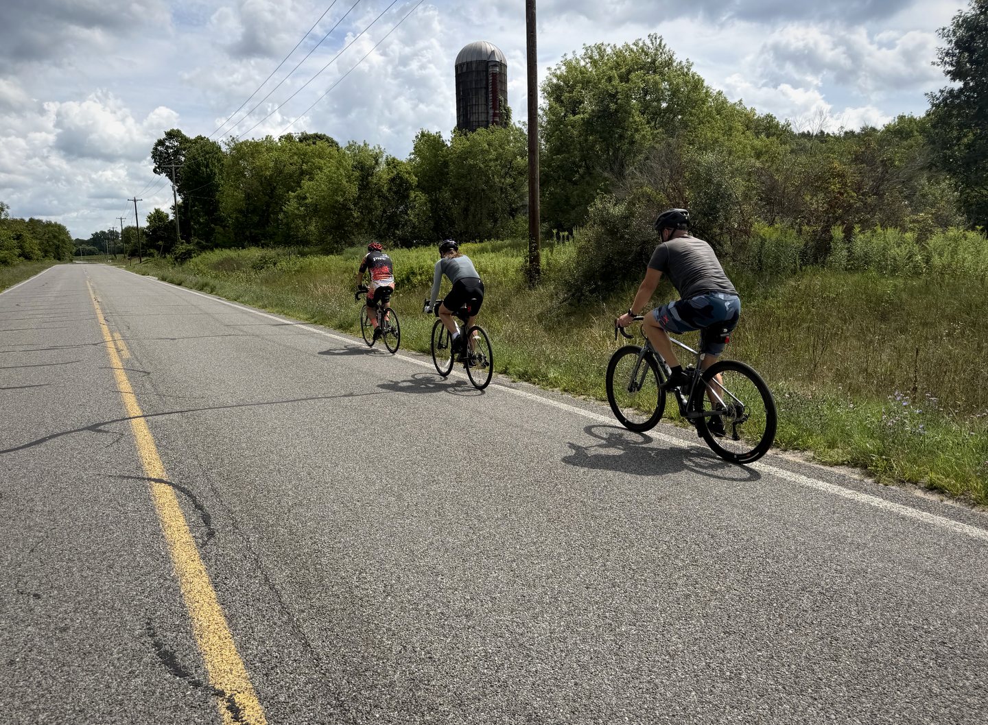 Three Pedal for Pride cyclists riding past a farm silo on a country road in 2026.