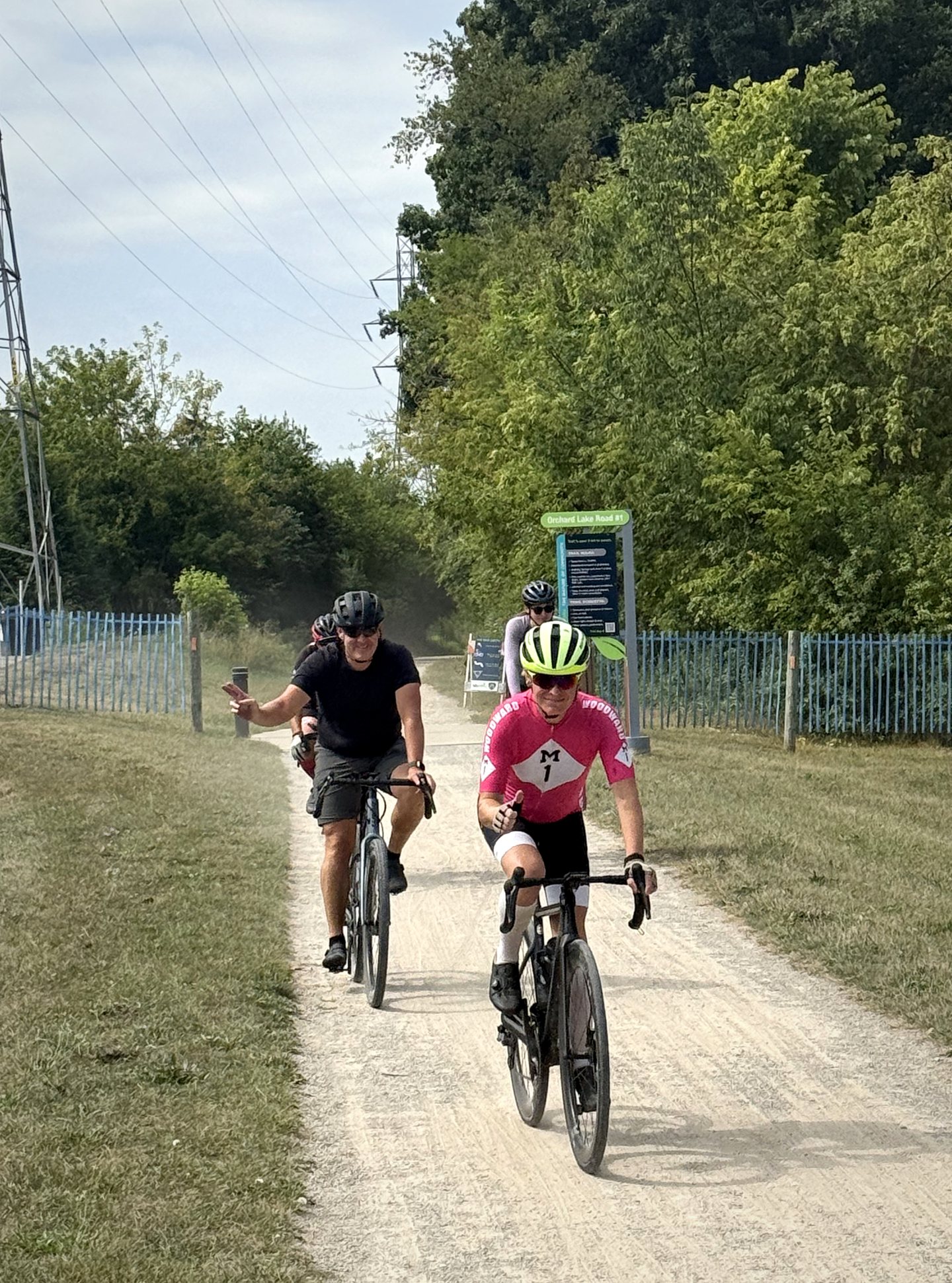 Three Pedal for Pride cyclists smiling as they ride toward the camera on a green trail in 2026.